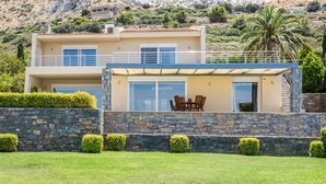 A two-story modern house with a stone facade, a balcony with outdoor furniture, and a view of a mountainous landscape.