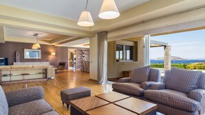 A modern living room with a sofa, ottoman, and coffee table. There is a kitchen area with a counter and cabinets, and a dining area with chairs. The room has large windows with a view of the sea.