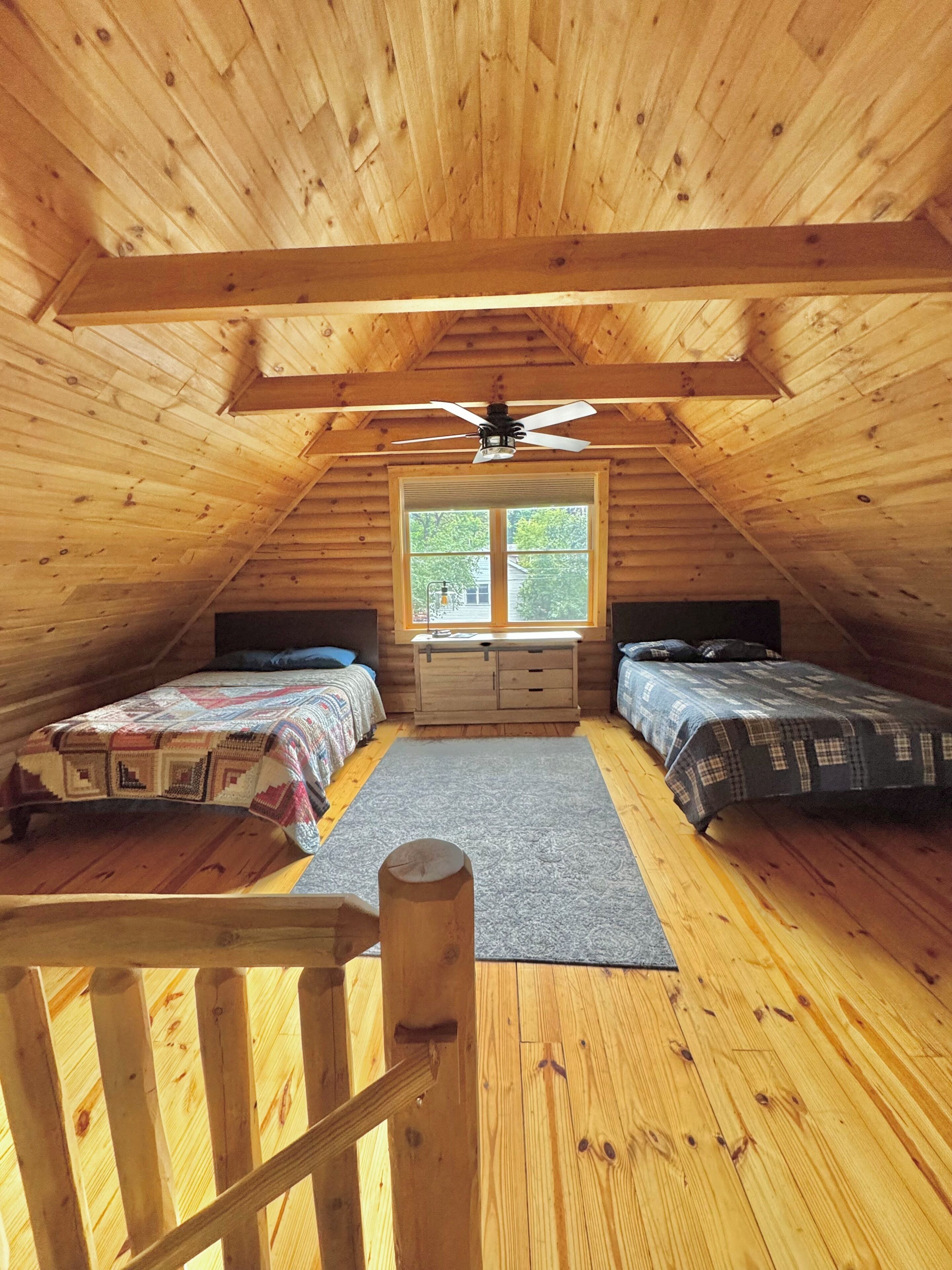 A cozy attic bedroom with two beds, a window, and a ceiling fan.
