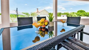 A glass-top table with wicker chairs, a fruit bowl, and a drink on a balcony with a clear view of the sky and greenery. - Vacation home Anić by Interhome (Novigrad (Zadar))
