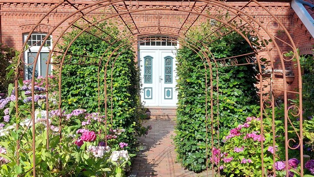 Un edificio in mattoni con tetto in tegole rosse, una porta bianca con infissi verdi e un vialetto in mattoni che conduce all'ingresso.