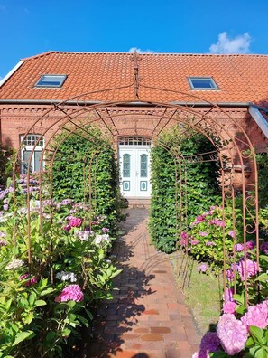 A brick building with a red-tiled roof, a central doorway, and a garden with a metal archway leading to the entrance. - To complete (Niedersachsen)