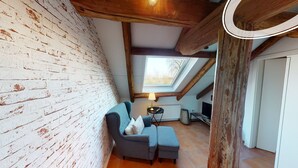 Una stanza in stile loft con travi a vista, soffitto inclinato e lucernario. Si vede un divano blu con un cuscino e un tavolino con una lampada.