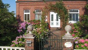 Una casa in mattoni con tetto in tegole rosse, una porta bianca e un giardino con fiori rosa e un cancello in metallo.