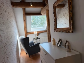 A room with a brick wall, a wooden ceiling beam, a window with a view of greenery, a black chair, a white dresser, and a mirror with an ornate frame. - To complete (Niedersachsen)