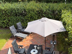 An outdoor patio with a brick floor, surrounded by greenery, featuring a table with chairs and a large umbrella. - To complete (Niedersachsen)