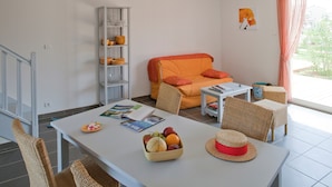 A living room with a white table, wicker chairs, a sofa with orange cushions, a bookshelf, and a window with curtains.