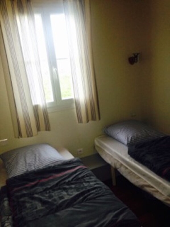 Quarto pequeno e pouco iluminado com duas camas de solteiro, uma janela com cortinas e luminária fixada na parede.