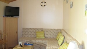 A cozy room with a wooden ceiling, a television mounted on a cabinet, a patterned sofa with cushions, a small table with a basket and books, and a wall-mounted light fixture. - wooden chalet in the gardens of St Pierre (Saint-Pierre-D'oléron)