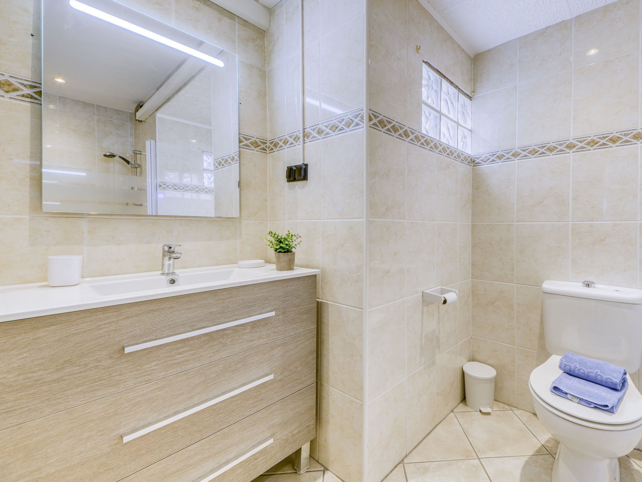 A modern bathroom with a beige tiled wall, a white sink with a mirror, a toilet, and a small plant on the vanity.