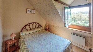 A bedroom with a bed, a nightstand, a lamp, a window with a view of the outdoors, and a wall-mounted heater.