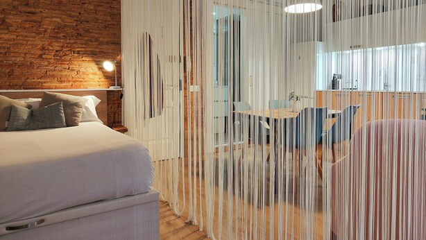 Une chambre moderne avec un mur en briques, un lit avec des oreillers, une table de chevet avec une lampe et un séparateur de pièce agrémenté d’éléments suspendus.