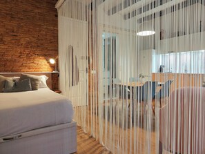 Ein modernes Schlafzimmer mit Backsteinwand, einem Bett mit Kissen, einem Nachttisch mit Lampe und einem Raumteiler mit hängenden Elementen.