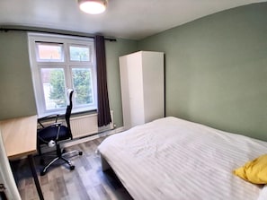 Un dormitorio con cama, escritorio con silla, radiador y ventana con cortinas.