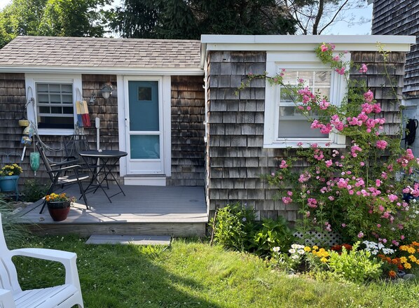 A small house with a porch, a door, and a window. There is a garden with various plants and flowers.