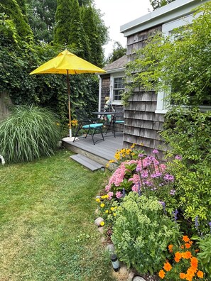 A backyard with a wooden deck, outdoor furniture, and a variety of flowering plants. - Cozy Cottage Old Fashioned Charm (Rockport)
