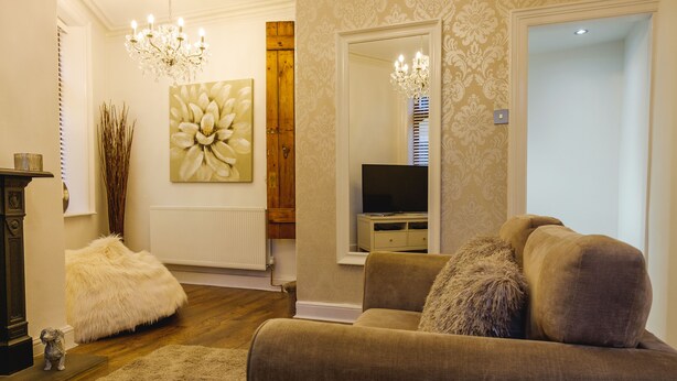 A living room with a brown sofa, a fluffy white pouf, a wooden door, and a chandelier. - St John's Cottage - Simple2let Serviced Apartments (Halifax)