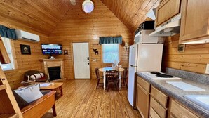 A cozy cabin interior with a wooden floor, a dining table with chairs, a kitchen area with a refrigerator, and a fireplace. - Ravine- Firepit, Pet Friendly, Cozy Cabin, WIFI, Pond, Gas Fireplace, off Blue Ridge Parkway (Laurel Springs)