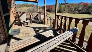 A wooden porch with a swing, outdoor seating, and a view of a forested landscape. - Ravine- Firepit, Pet Friendly, Cozy Cabin, WIFI, Pond, Gas Fireplace, off Blue Ridge Parkway (Laurel Springs)