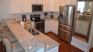 A kitchen with white cabinets, stainless steel appliances, granite countertops, and a bar area with bar stools. - Barefoot 'N Salty - Walk to Beach in Surfside Beach (Surfside Beach)