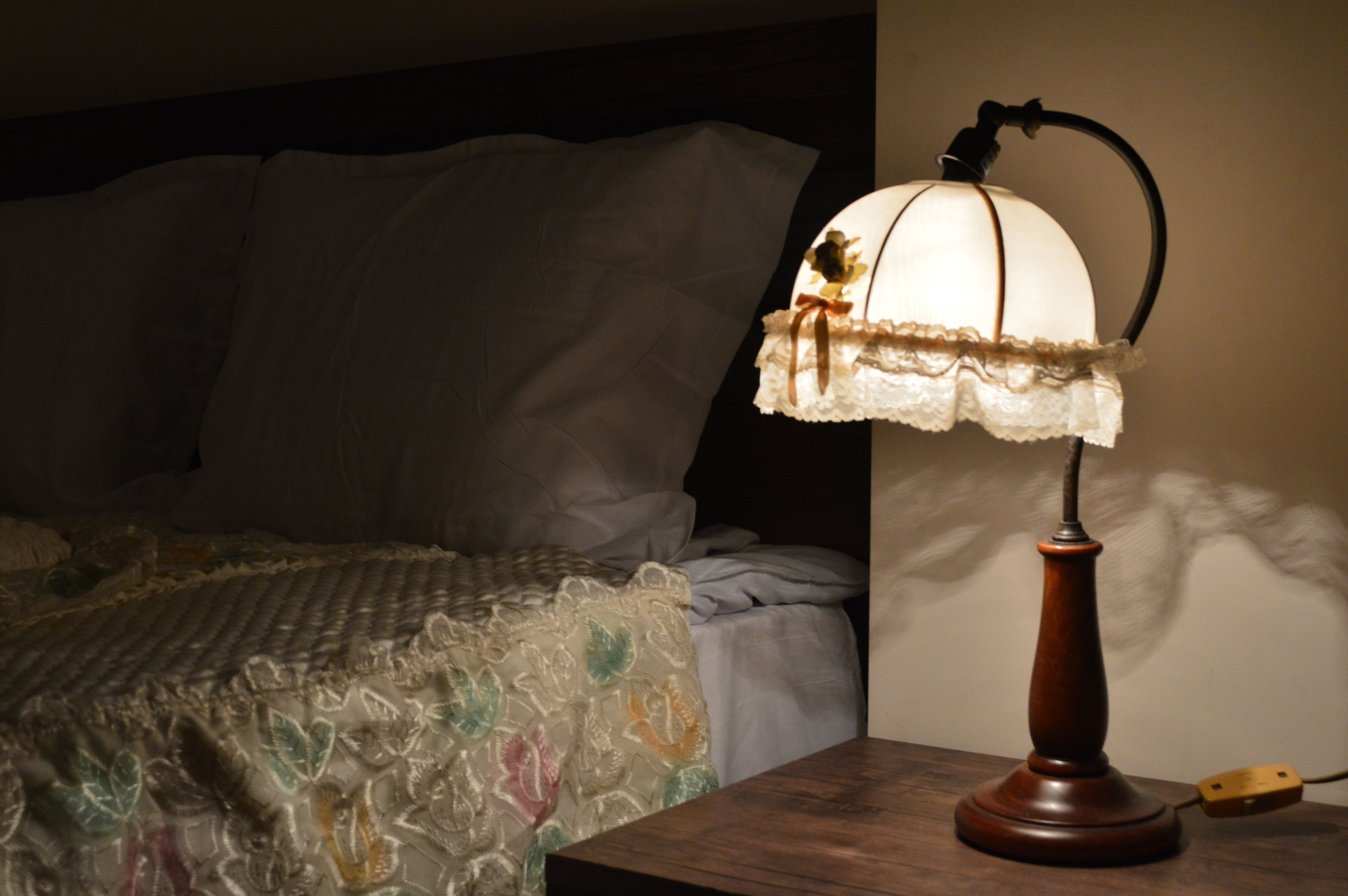 A bed with a floral-patterned bedspread and a vintage-style table lamp with a lace shade on a wooden nightstand.