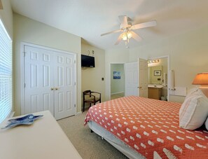 A bedroom with a bed, a desk, a chair, a ceiling fan, a TV, and a bathroom mirror. - Fenway Park - Second Row in Surfside Beach (Surfside Beach)