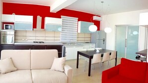 A modern kitchen with white cabinets, a red accent wall, and a dining area with a red chair and a white sofa.