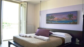 A bedroom with a bed, a painting of a beach scene, and a view of the outdoors through a sliding door.
