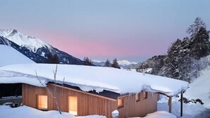 Eine Holzhütte mit schneebedecktem Dach, umgeben von Schnee und Bergen, mit sichtbarer Tür und Fenstern.