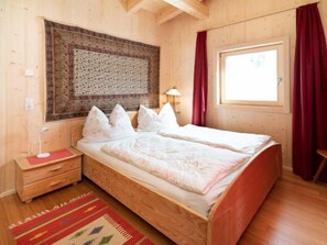 Ein Zimmer mit Holzboden, einem Bett, einem Nachttisch, einer Lampe und einem Fenster mit Vorhängen.