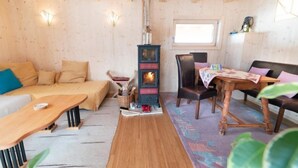 Ein gemütliches Zimmer mit Holzbalkendecke, einem zentralen Kamin, einer Couch, einem Esstisch und einem Fenster.