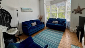 A living room with two blue sofas, a black wicker chair, a wooden floor, and a large window with blinds. - Great location, close to Ventnor beach and town. Perfect for families. (Isle of Wight)