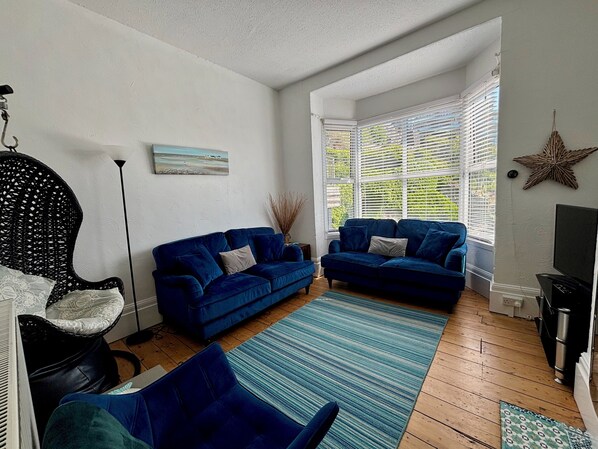 A living room with two blue sofas, a black wicker chair, a wooden floor, and a large window with blinds.