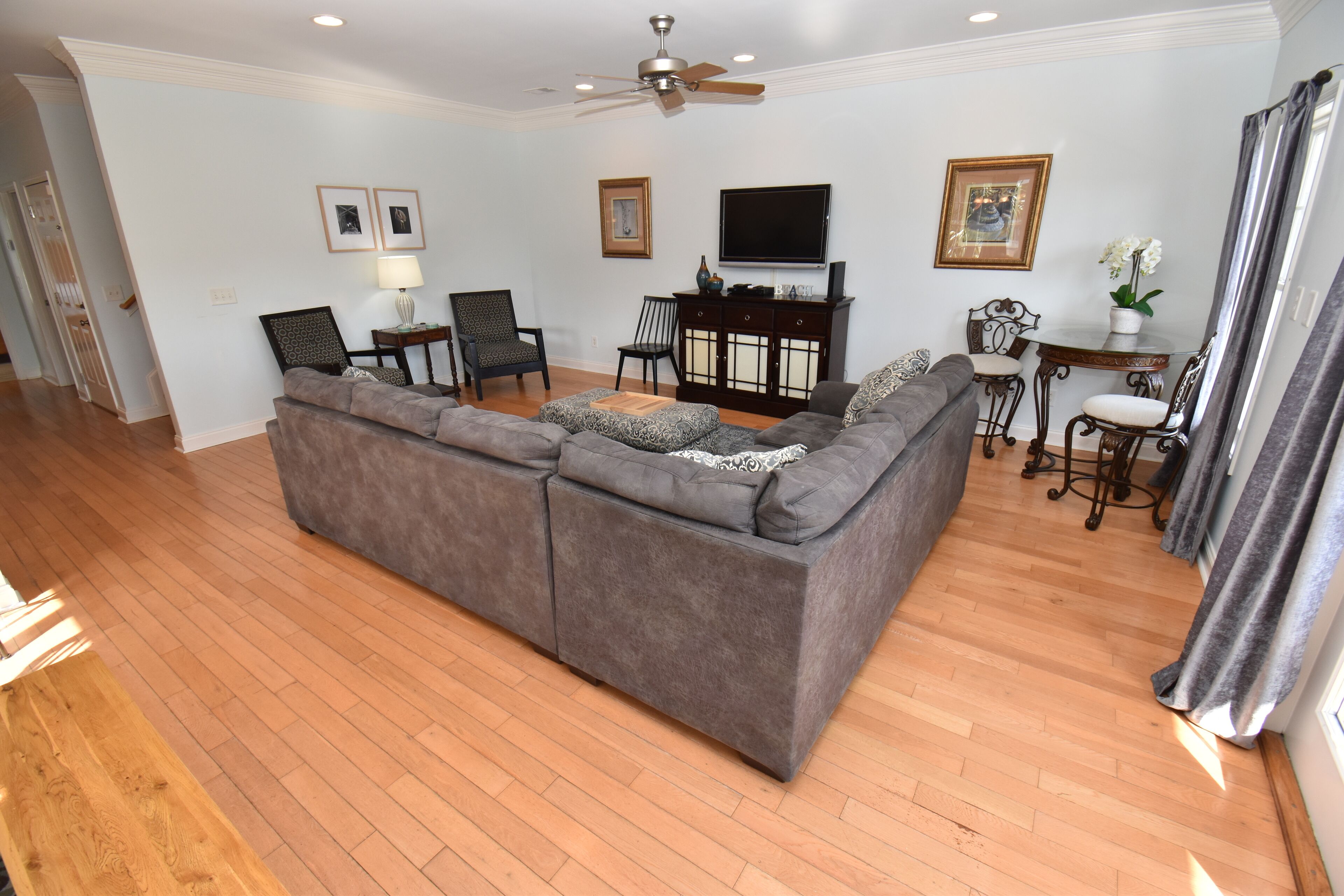 A living room with a gray sectional sofa, a flat-screen TV, a ceiling fan, and a dining area with a table and chairs.