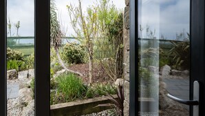 Une vue à travers une porte vitrée d’un jardin avec un mur de pierre, un banc en bois et un sentier.