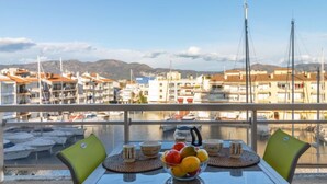 Een tafel met een fruitschaal, kopjes en een waterkoker op een balkon met uitzicht op een jachthaven met boten en gebouwen.