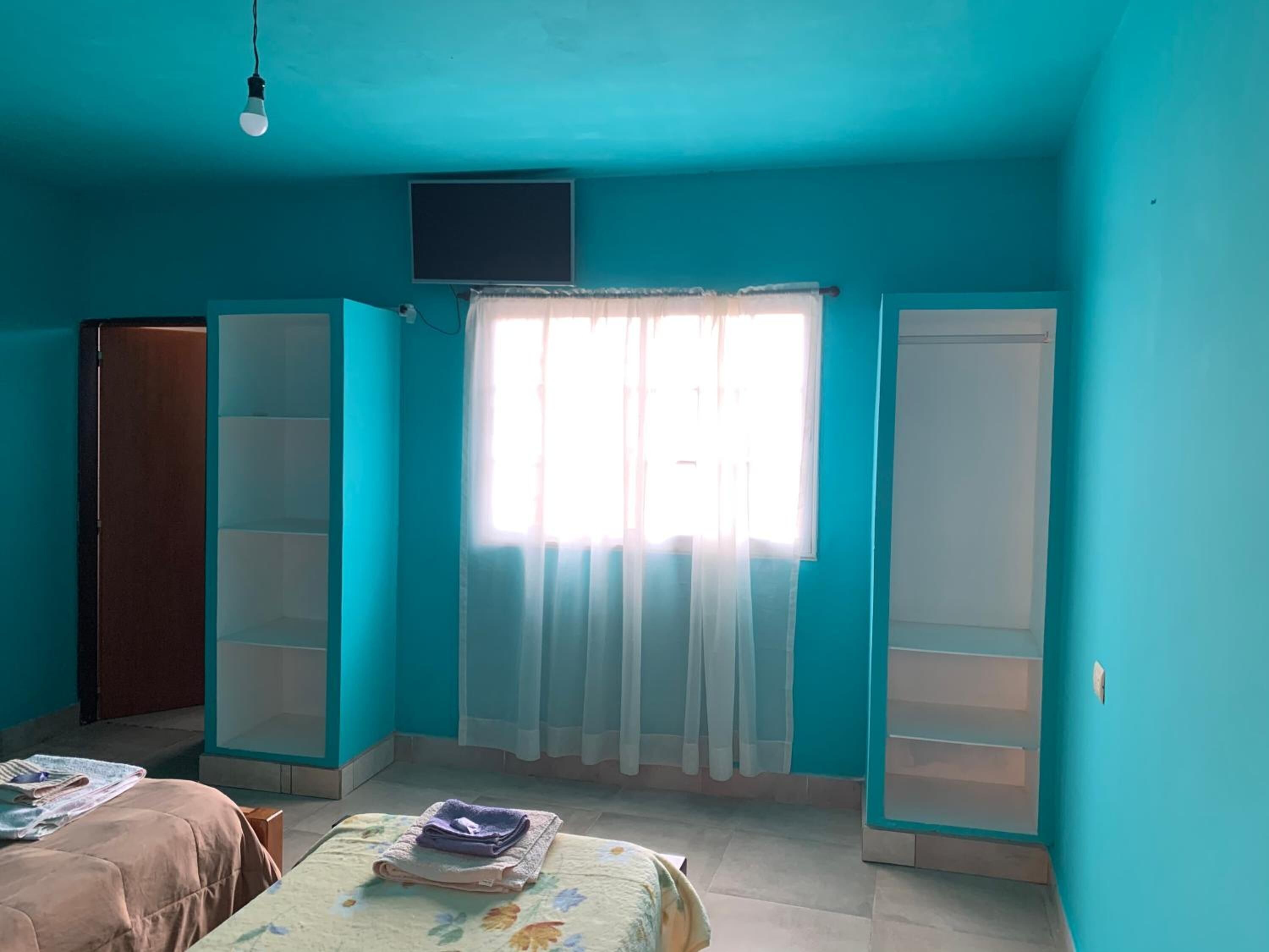 A room with teal walls, a bed with a floral bedspread, a window with sheer curtains, and a closed wardrobe.