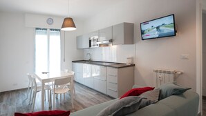 Una cocina moderna con gabinetes blancos, un comedor con mesa y sillas blancas, y una sala de estar con sofá gris y almohadas rojas.