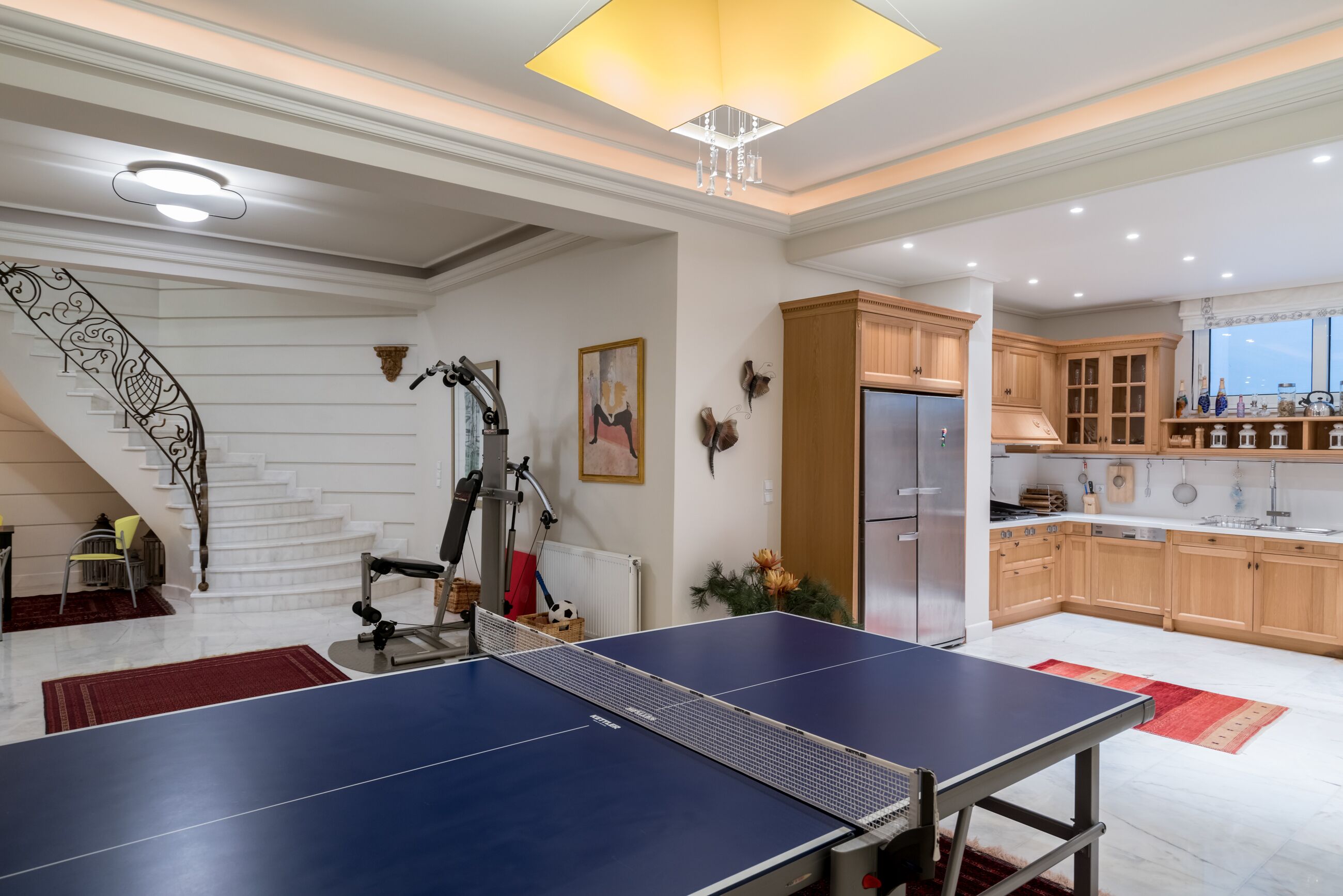 A spacious living area with a ping pong table, a staircase with decorative railings, and a kitchen with wooden cabinets and a large window.