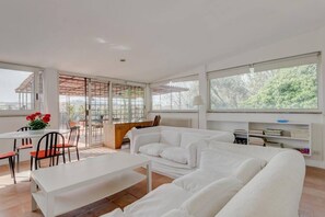 Uma sala de estar moderna com um sofá branco, uma mesa de jantar e cadeiras. Há uma janela grande com vista para vegetação.