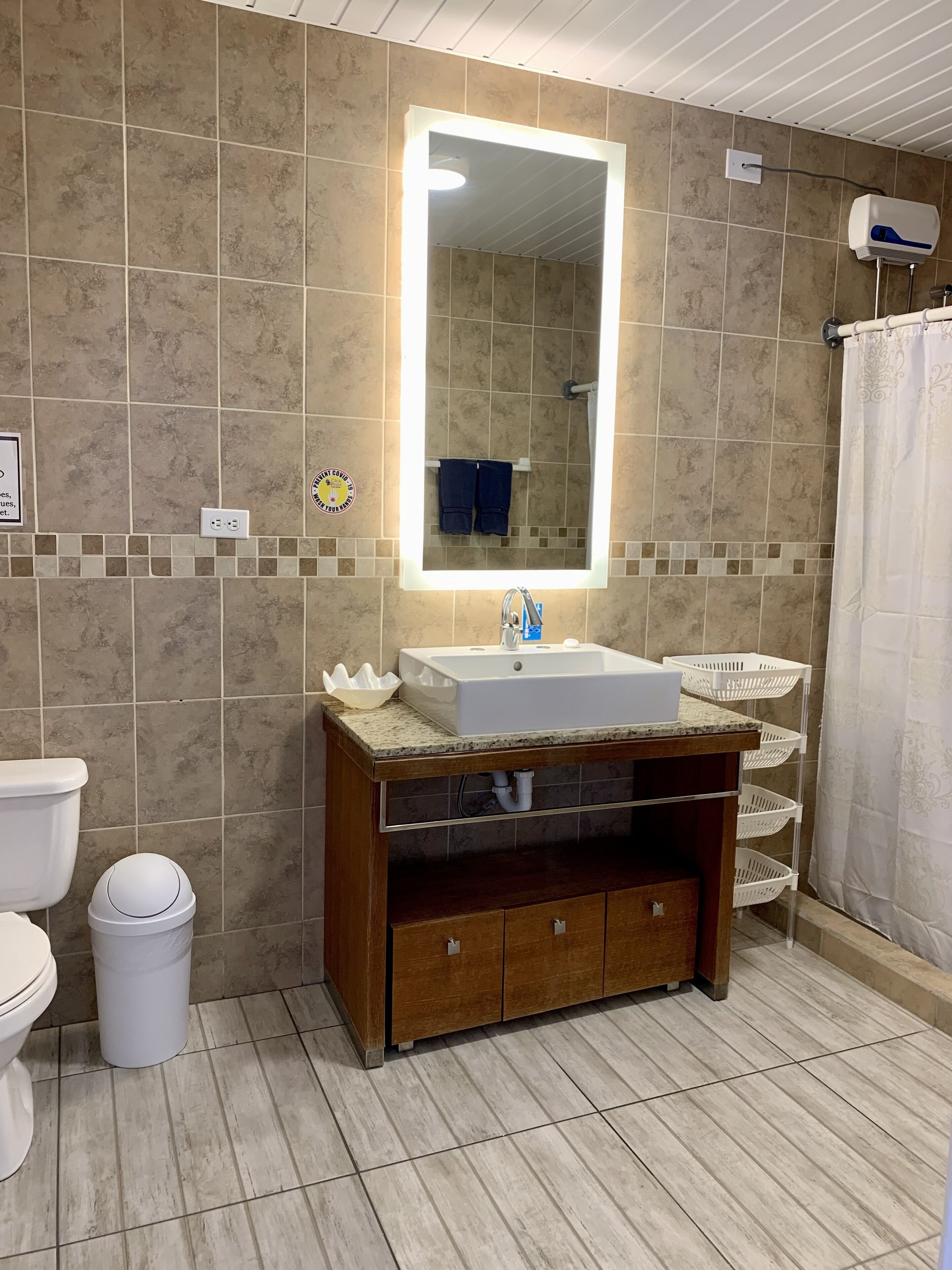 A bathroom with a sink, mirror, and tiled walls. There is a toilet, a trash can, and a shower curtain.