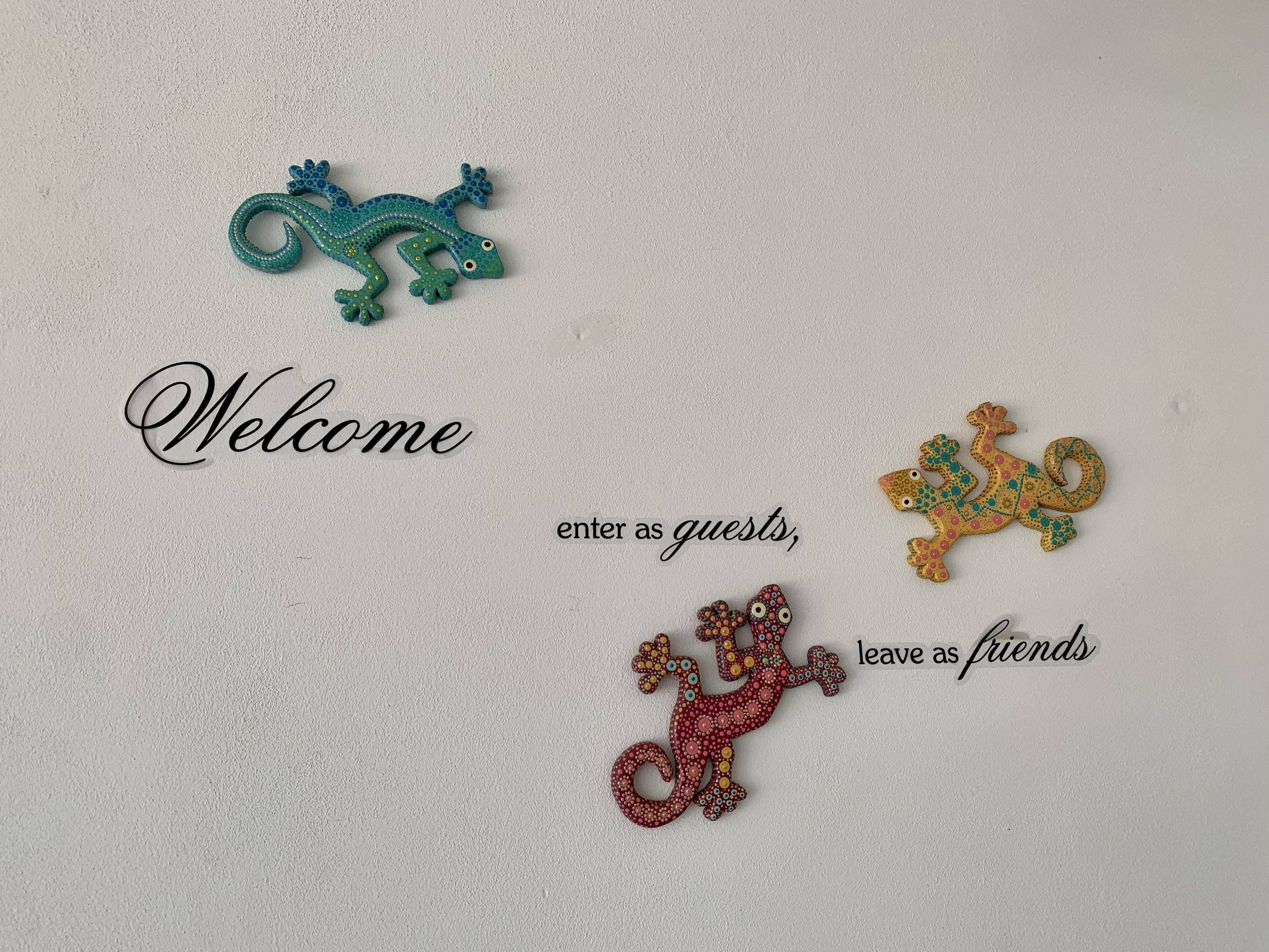 Een verwelkomingsbord met een decoratieve hagedis en de tekst 'kom binnen als gast, vertrek als vriend' op een gestructureerde muur.