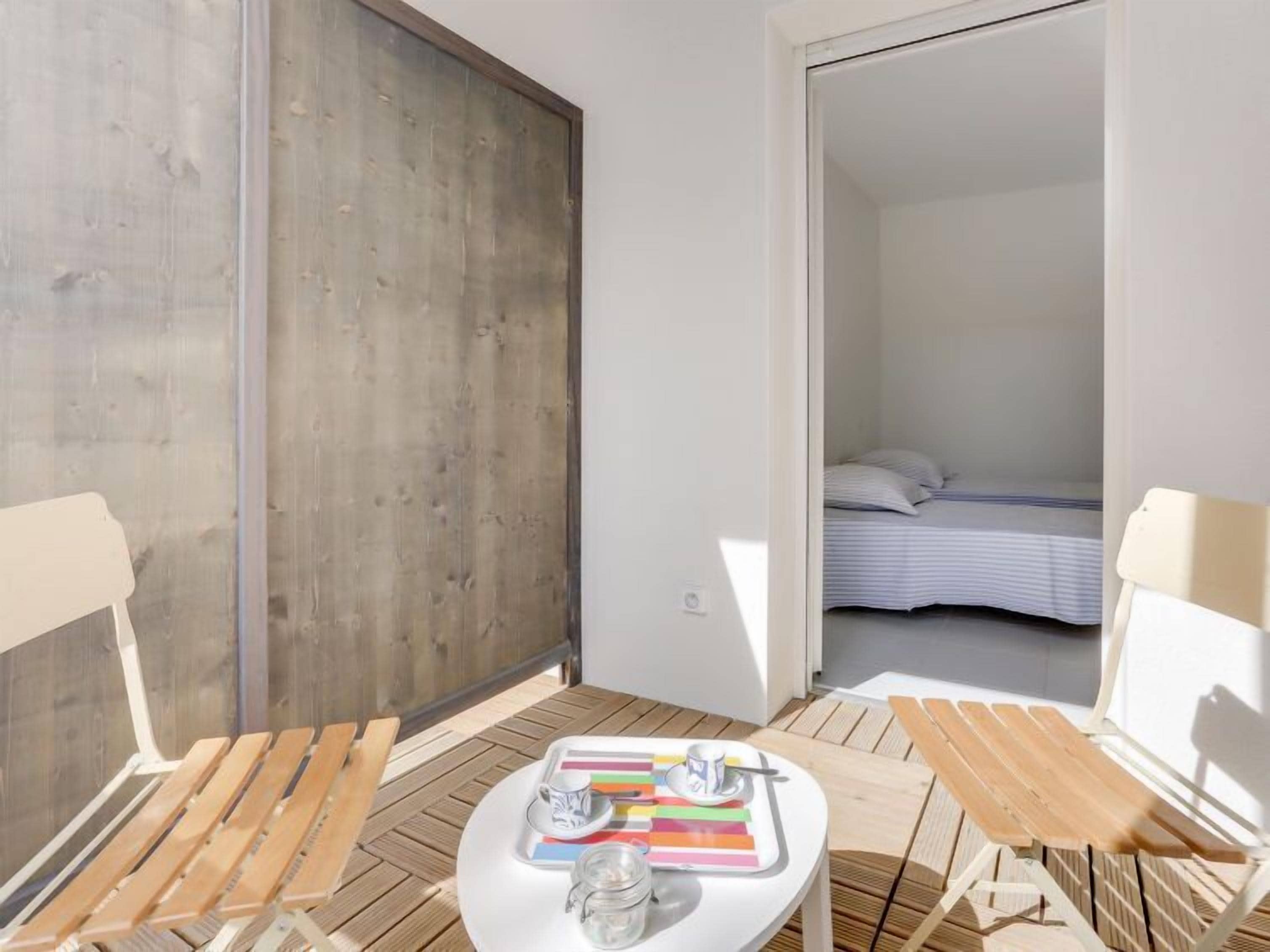 Een kleine, lichte kamer met een houten vloer, een bed en een tafel met een dienblad met boeken en een kop.