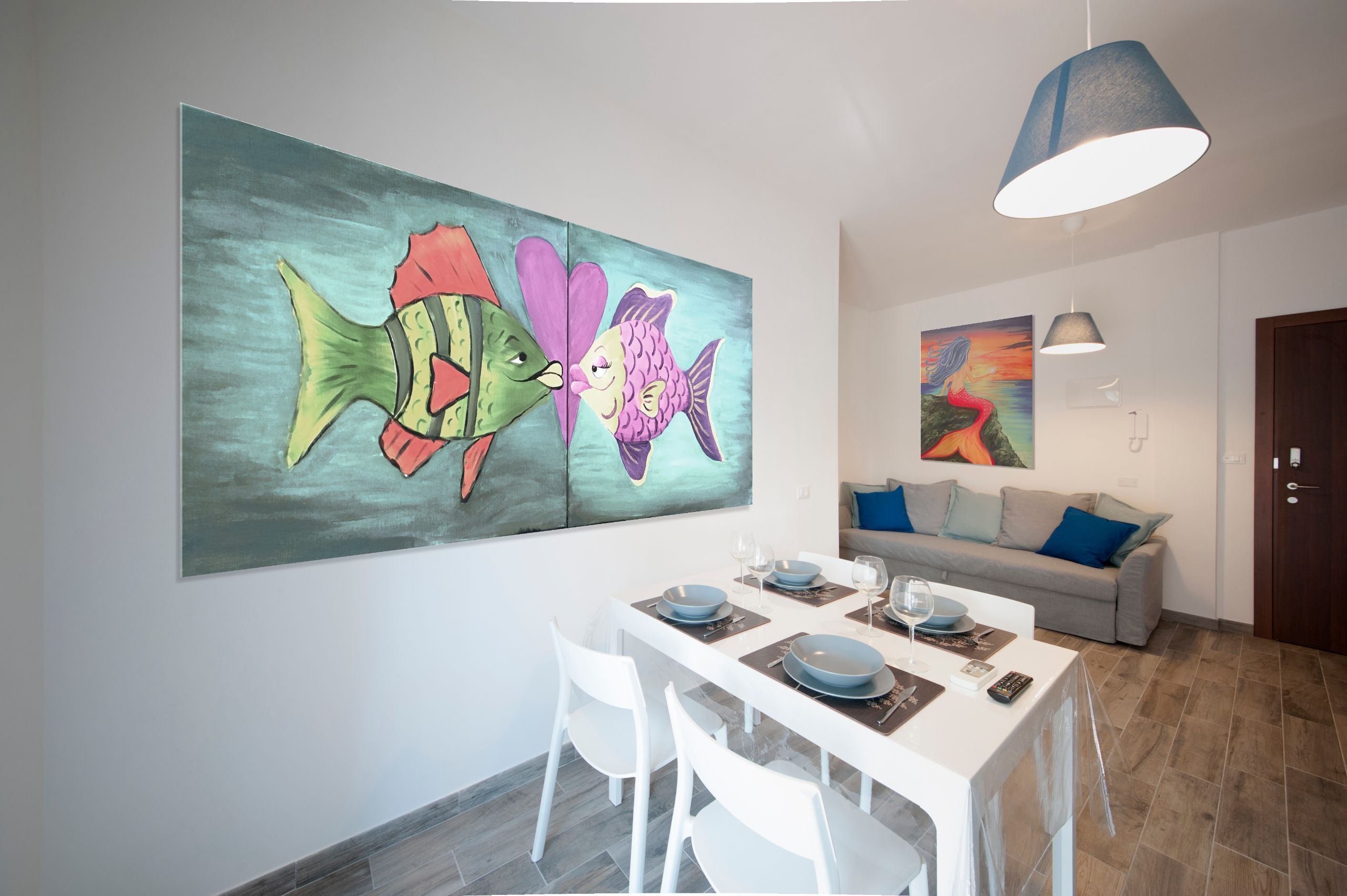 Ein Essbereich mit einem weißen Tisch, gedeckt für vier Personen, einem großen farbenfrohen Gemälde von einem Fisch an der Wand und einem grauen Sofa mit blauen Kissen im Hintergrund.