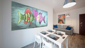 Un comedor con una mesa blanca dispuesta para cuatro personas, un cuadro grande de un pez colorido en la pared, y un sofá gris con cojines azules al fondo.