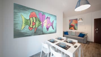 Ein Essbereich mit einem weißen Tisch, gedeckt für vier Personen, einem großen farbenfrohen Gemälde von einem Fisch an der Wand und einem grauen Sofa mit blauen Kissen im Hintergrund.