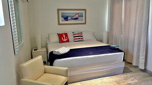 A neatly arranged hotel room with a bed, a chair, a nightstand with a lamp, and a framed picture on the wall. - Tu Casita en Aruba (CRISTINA KING) (Noord)