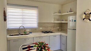 A compact kitchen with a sink, stove, and a refrigerator. There are shelves above the stove holding decorative items and a clock with "Welcome Aruba" on it. - Tu Casita en Aruba (CRISTINA KING) (Noord)