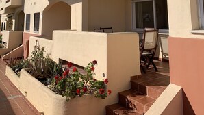 Un immeuble à plusieurs étages, doté d’un toit plat, de balcons et d’un petit jardin agrémenté de fleurs rouges.