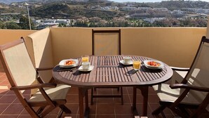 Une table ronde en bois, sur laquelle sont disposées deux assiettes de nourriture et deux tasses de boissons, se trouve sur un balcon offrant une vue sur un paysage urbain et des montagnes en arrière-plan.